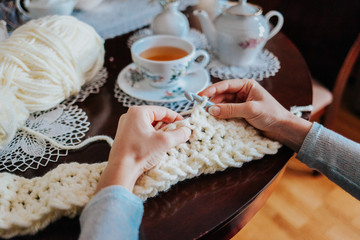 Woman's hands crocheting with white wool on the table with a cup of tea