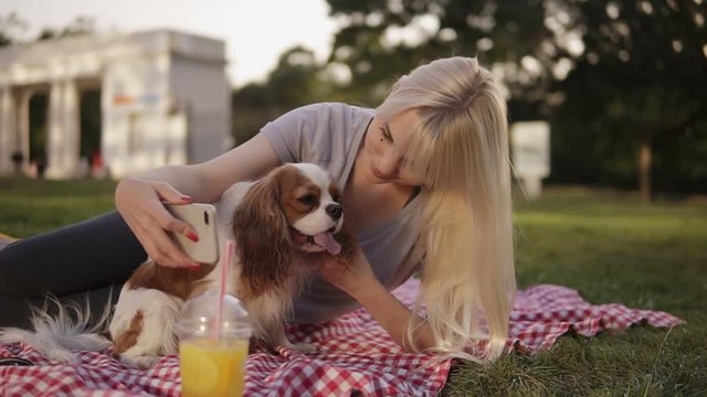 Blonde long haired woman laying on on a plaid in the park and trying to make a selfie photo with her spaniel using a smartphone. Green grass, trees on the background