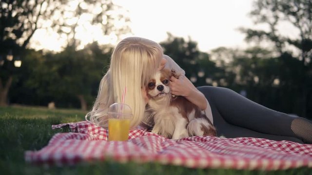 Beautiful caucasian blonde woman with her dogs in park, relaxed on green grass on a litter - caress and cuddling her loving pet - cavalier king charles spaniel