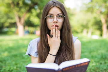 Fototapeta premium Shocked woman sitting in park while holding book and looking at the camera