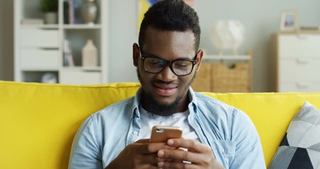 Portrait shot of the cheerful young African American man taping while texting on the smartphone device in the living room. - Powered by Adobe