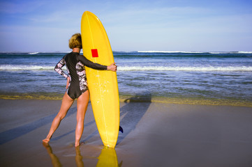 back view of young attractive and sporty surfer girl in cool swimsuit at the beach holding yellow surf board looking at the blue sea n in summer surfing holidays