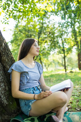 Side view of pleased brunette woman in eyeglasses sitting on the grass under tree and reading book in park