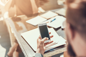 Close up male hand holding contemporary cellphone in hand. He looking at screen of appliance. Different documents locating on table