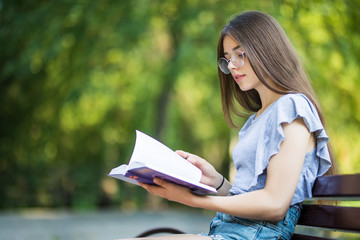 Side view of pleased woman in eyeglasses sitting on bench and reading book in park