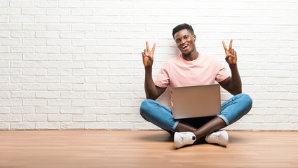 Afro american man sitting on the floor with his laptop smiling and showing victory sign