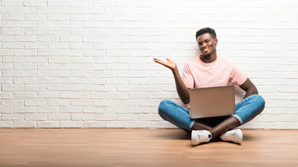 Afro american man sitting on the floor with his laptop holding copyspace imaginary on the palm