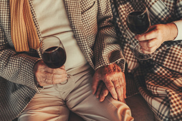 Top view of wrinkled female and male hands holding glasses with wine. Couple sitting on couch