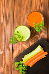 Carrot and celery juice in glasses with cut fresh vegetables and parsley leaves on kitchen board on wooden background.