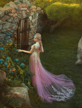 cute princess wearing long pink dress with trailer and is walking in the garden and touching flowers in soft sun lights. wonderful elegant girl elf with blond fair wavy hair and gold crown. art photo