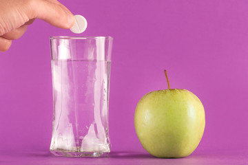 Hand throwing effervescent tablet vitamin mineral supplement into glass of water and green apple isolated on purple background. Health and medical concept