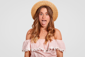 Funny beautiful comic woman shows tongue, foolishes with friends, wears fashionable blouse, straw summer hat, sees something amazing, isolated over white background. People and emotions concept