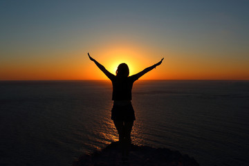 Fototapeta premium young girl is facing the sun on the mountain and looking at the sea, hands raised up and to the side