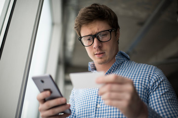 Handsome man holding business card and mobile phone in office. Well dressed businessman wearing stylish glasses researching information online. Guy call a taxi indoors.