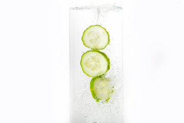 Slices of cucumber fall into clear clear water. There are bubbles in the water. On a white background. Close-up.