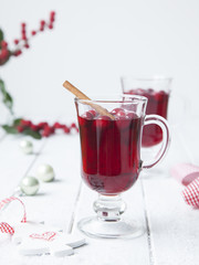 Hot red berry winter drink in two glasses on white wooden table with Christmas tree toys on background.