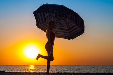 Silhouette of a girl in a swimsuit with a beach umbrella on the coast at sunrise or sunset.