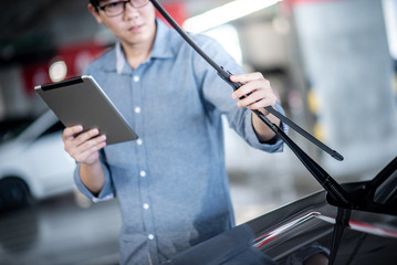 Young Asian auto mechanic holding digital tablet checking windshield wiper in auto service garage. Mechanical maintenance engineer working in automotive industry. Automobile servicing and repair