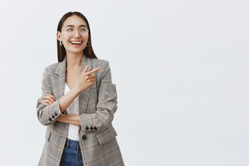 Horizontal shot of stylish feminine girlfriend in glasses and trendy earrings, laughing from pleasure and happiness while looking and pointing at upper right corner, being amused over gray wall