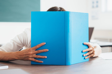 Student girl covering her face behind a book in classroom.