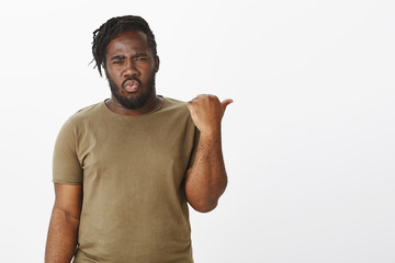 Portrait of shocked questioned attractive african man in casual outfit, folding lips and frowning, pointing right with thumb, asking question about something interesting, standing over gray background
