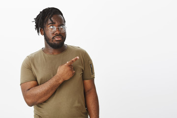 Studio shot of intense suspicious attractive dark-skinned man in casual t-shirt, staring and indicating at upper right corner, being unsure or displeased, standing in doubts over gray background