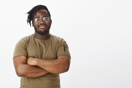 Smart happy and confident african-american in stylish transparent glasses, holding hands crossed, standing in self-assured pose, smiling broadly at camera, posing for advertisement over grey wall