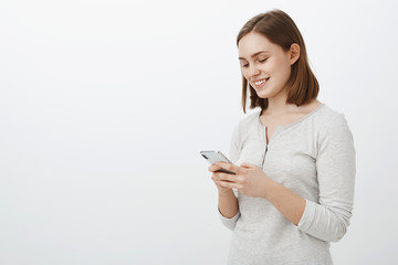 Fototapeta premium Waist-up shot of sociable carefree female internet geek smiling joyfully while having amusing chat in network with best friend from online game holding smartphone and smiling at cellphone screen
