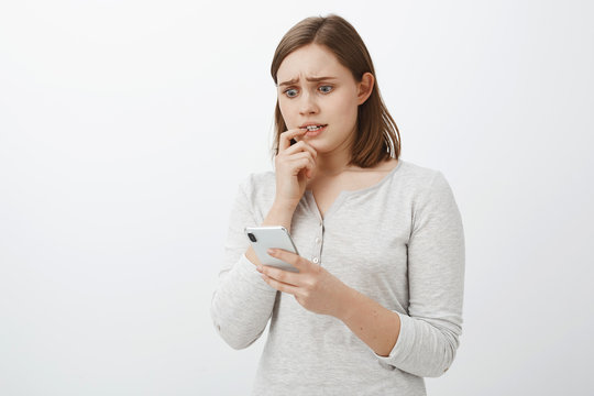 Girl feeling nervous sending money via app to wrong person biting fingernail frowning staring anxiously at smartphone screen being shocked and scared of bad consequences of action over gray wall