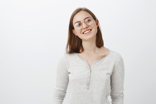 Waist-up shot of smart creative charming young woman with short cute haircut tilting head joyfully smiling broadly wearing glasses and casual white blouse posing happily over grey background