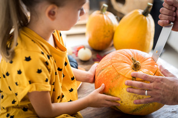Cute little girl sitting on kitchen table, helping her father to carve large pumpkin, smiling....
