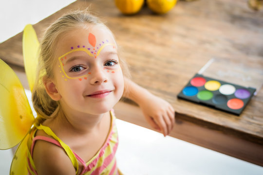 Cute little girl with face paint sitting at a table, looking at camera smiling. Halloween party or carnival family lifestyle background. Face painting and dressing up.