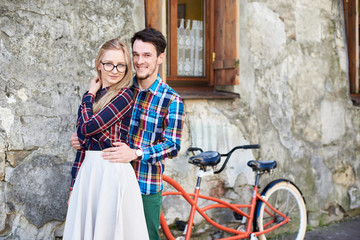 Fototapeta na wymiar Portrait of young attractive tourist couple, bearded man and blond woman standing together, smiling and looking to the camera at modern tandem bicycle on empty pavement at cracked wall of old building