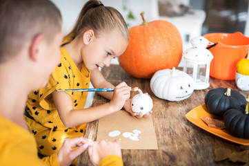 Cute young girl sitting at a table, decorating little white pumpkins with her mother. Halloween holiday and family lifestyle background.