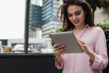 Smiling woman reading funny articles on tablet white standing outside and waiting for her friend. Hipster girl watching video on touch pad using wireless internet. Copy space for you logo or design