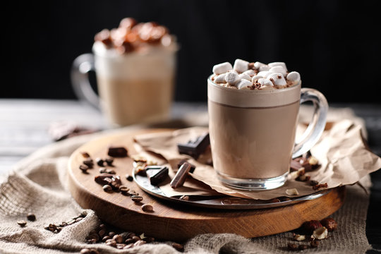 Composition with two glass of cappuccino topped with marshmallow and served with coffee beans and chocolate on wooden chopping board over black background