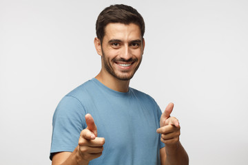 Hey you! Young man in blue t-shirt pointing to camera with fingers isolated on gray background