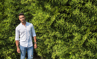 Attractive handsome young man in city park, standing against big bush, in a summer day, wearing jeans and white shirt