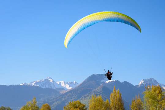 Paraglider in Annecy, Apls, France. Unidentified man flies with paraglide above trees with snow top rocky cliff mountains in background. Descend at sunset at Annecy lake, french Apls, France.