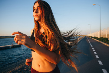 Gorgeous young girl with long brown hair in a sports top and tights runs on the road along the reservoir on the sunset