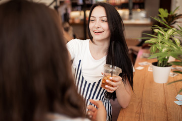 Two beautiful youthful smiling girls with dark hair,dressed in casual outfit,stand opposite each other and chat in a cozy coffee shop.