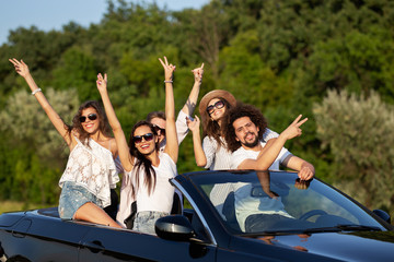 Happy beautiful young  dark-haired girls and guys in sunglasses smile in a black cabriolet on the road holding their hands up on a sunny day.