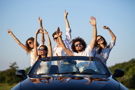 Happy young girls and guys in sunglasses are sitting in a black cabriolet on the road holding their hands up and smiling on a sunny day.