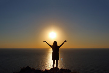 Fototapeta premium young girl is facing the sun on the mountain and looking at the sea, hands raised up and to the side