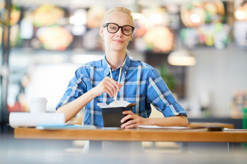 Fototapeta na wymiar Young blond casual woman in checkered shirt and eyeglasses looking at you while siting by table and having noodles from box