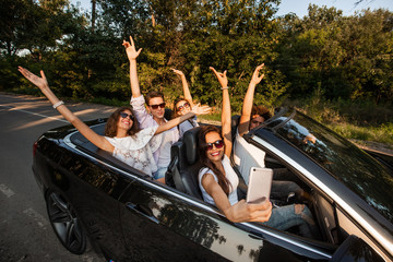 Group of young girls and guys are holding their hands up and making selfie in a black cabriolet on...