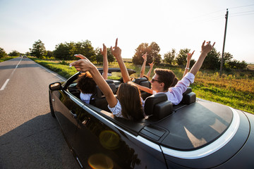 Black cabriolet is on the country road. Happy group of young girls and guys are sitting in the car...