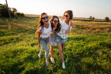 Three gorgeous young women in sunglasses dressed in the beautiful clothes stand in the field and smiling on a sunny day.