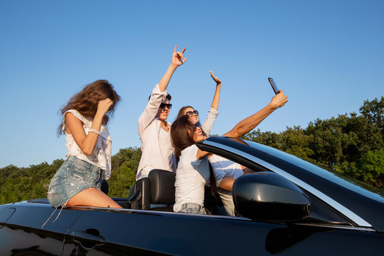 Joyful young girls and guys are sitting in a black cabriolet on the road and making selfie on a sunny day.