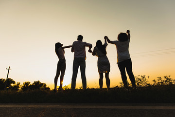 Company of young girls and guys are standing on the grass near the road on a summer day and holding their hands.